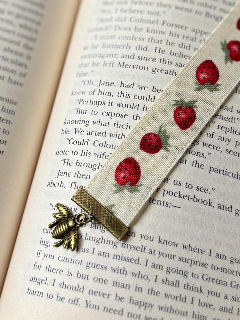 May include: A cream fabric bookmark featuring a strawberry pattern and a bronze bee charm. The bookmark is inserted into a book, with the text of the book visible. The bookmark is approximately 20 cm long.