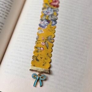 May include: A yellow floral fabric bookmark with a scalloped edge. It features a gold-coloured metal accent and a blue bow charm. The bookmark is in an open book.