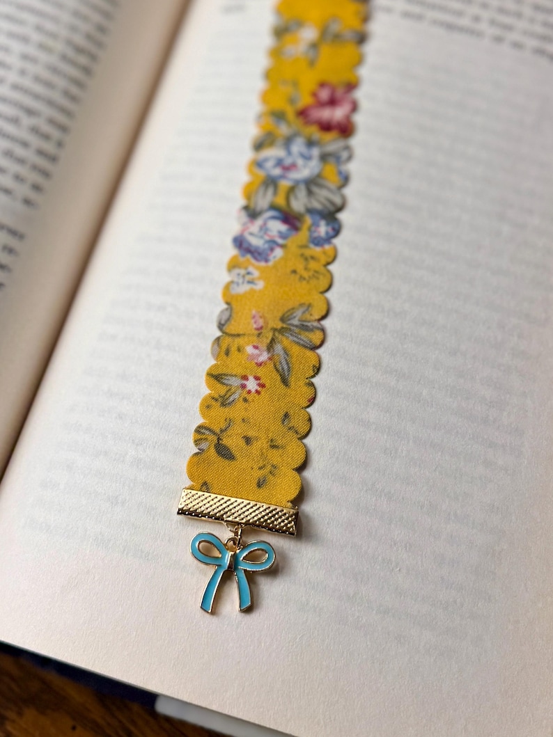 May include: A yellow floral fabric bookmark with a scalloped edge. It features a gold-coloured metal accent and a blue bow charm. The bookmark is in an open book.