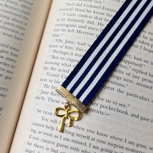 May include: A navy and white striped ribbon bookmark with a gold bow charm, placed in a book. The bookmark is a decorative accessory for readers.