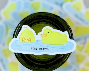 Puede incluir: Una pegatina de pato amarillo con el texto "stay weird". La pegatina presenta dos patos amarillos en una escena de agua azul. La pegatina está sobre una superficie verde oscuro, con otras pegatinas de patos en el fondo.