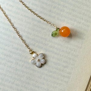 Op de afbeelding: Delicate goudkleurige kettingoorbellen. Eén oorbel heeft een witte bloemhanger met gouden accenten. De andere oorbel heeft een oranje kraal met een groene bladhanger. De oorbellen staan op een lichte achtergrond.