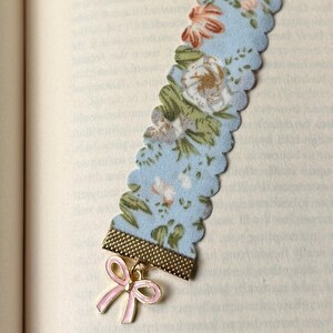 May include: A light blue floral fabric bookmark with a scalloped edge. The bookmark has a gold-coloured metal band and a pink bow charm. The fabric features a pattern of white, pink, and green flowers.