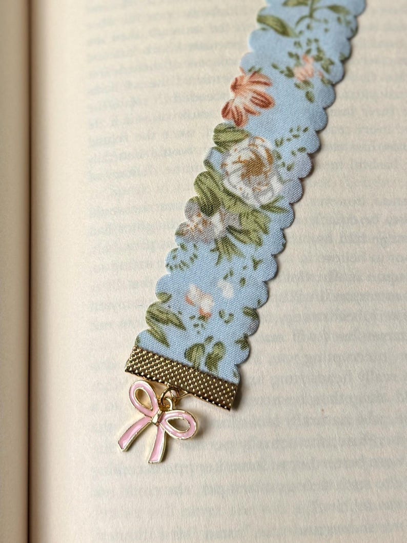 May include: A light blue floral fabric bookmark with a scalloped edge. The bookmark has a gold-coloured metal band and a pink bow charm. The fabric features a pattern of white, pink, and green flowers.