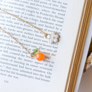 May include: A gold-toned chain necklace featuring two charms: a white five-petaled flower with a gold center and a bright orange spherical fruit charm topped with a small green leaf. The necklace rests on an open book page with printed text.