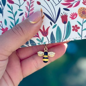 May include: A floral patterned wallet with a gold and rhinestone bee charm. The wallet features a white background with pink, red, and orange flowers and green leaves. The bee charm has yellow and black stripes.