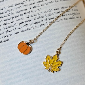 Könnte beinhalten: Zwei goldfarbene Halsketten mit Anhängern zum Thema Herbst. Ein Anhänger ist ein orangefarbener Kürbis, der andere ein gelbes Ahornblatt. Die Halsketten werden auf den offenen Seiten eines Buches präsentiert.