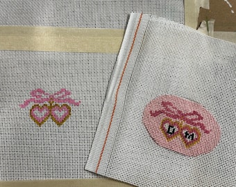 May include: Two pieces of needlepoint canvas with pink and gold heart designs. One canvas features two hearts with a bow, and the other has a pink oval with a heart design and the letters "D YA".