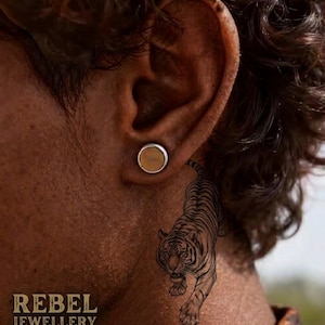 May include: A close-up of an ear with a circular ear plug. The plug has a silver rim and a light brown centre. A detailed tattoo of a tiger is on the person's neck. The text "REBEL JEWELLERY" is at the bottom.