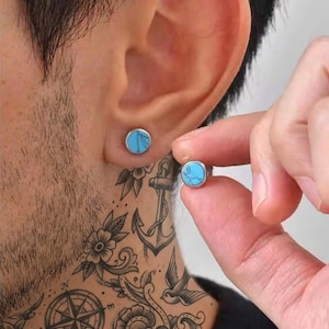Pode incluir: Brincos redondos com uma pedra turquesa brilhante em metal prateado. Um brinco está na orelha e o outro é segurado por uma mão. Os brincos têm um design simples e minimalista.