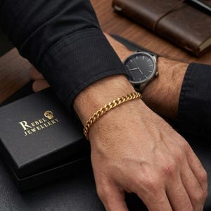 Pode incluir: Uma pulseira de corrente dourada no pulso, combinada com um relógio preto. A pulseira é exibida com uma caixa preta que tem o texto "REBEL JEWELLERY" e um logotipo de caveira. A pessoa está usando uma camisa preta de manga comprida.