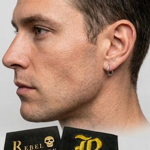 May include: A silver hoop earring is worn on the ear of a person. The image also shows two black pouches with gold text that reads "Rebel Jewellery" and a gold "R" logo.