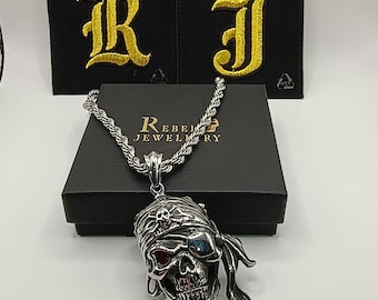 May include: A silver skull pendant necklace with a pirate theme. The skull has a bandana, an eye patch, and a detailed design. The necklace is on a black box with the text "Rebel Jewellery". Two black patches with gold embroidered letters are in the background.