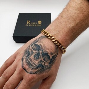 May include: A gold curb chain bracelet. The bracelet is shown on a hand with a skull tattoo. A black box with the text "Rebel Jewellery" is in the background.