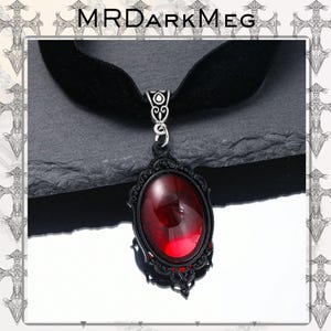 May include: Black velvet choker necklace with a gothic-style pendant. The pendant features a large, oval, deep red gemstone set in a black filigree frame. The necklace is a dark, elegant accessory.