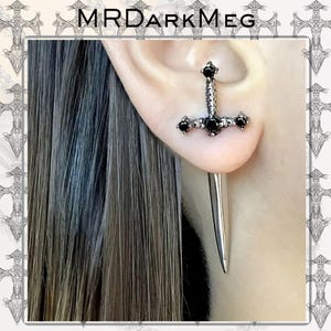 May include: A silver sword-shaped earring with black gemstone accents. The earring is worn in the ear, with the sword's blade extending downward. The earring has a gothic or fantasy theme, with the text "MRDARKMEG" at the top.
