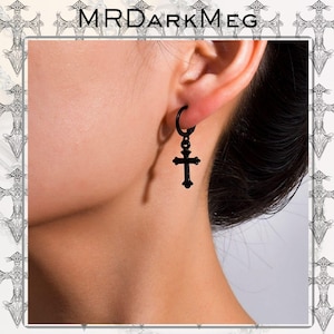 Puede incluir: Pendientes de cruz negros. Los pendientes presentan un colgante de cruz negro que cuelga de un aro negro. La imagen muestra el pendiente en la oreja. La marca "MRDARKMEG" está en la parte superior de la imagen.
