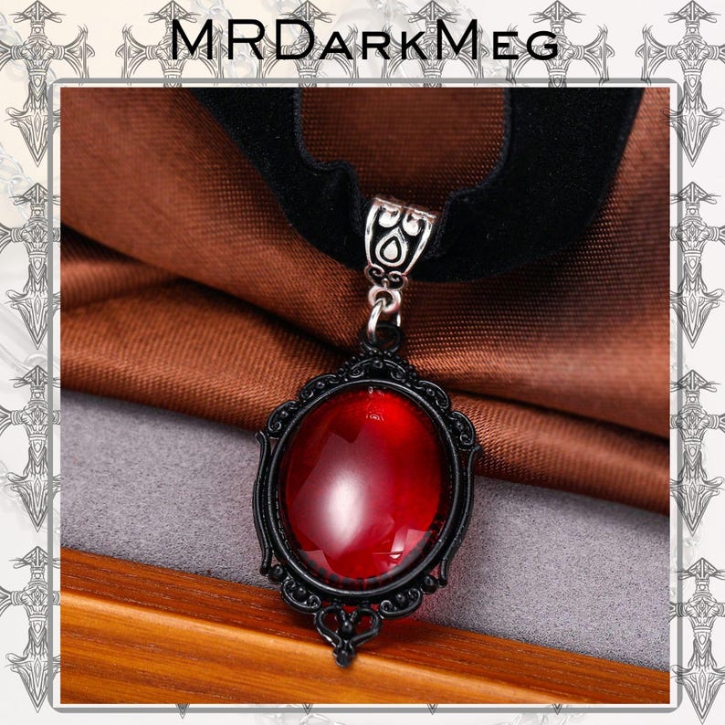 May include: Black velvet choker necklace with a large, oval, red gemstone pendant. The pendant is set in a black filigree frame. The necklace has a silver-toned clasp and the text "MRDARKMEG" is at the top.