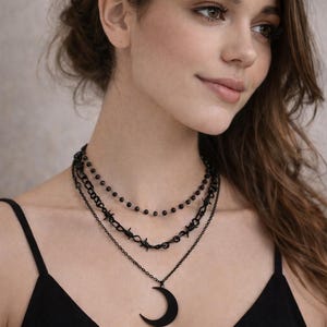 Könnte beinhalten: Ein mehrschichtiges schwarzes Halsketten-Set mit einem Halbmond-Anhänger. Das Set enthält eine Kette mit kleinen Perlen, eine Kettengliederkette und ein Stacheldraht-Design. Der Schmuck wird von einer Person getragen, die ein schwarzes Oberteil trägt.