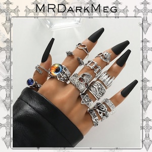 Puede incluir: Una mano adornada con múltiples anillos plateados con diversos diseños, incluyendo calaveras y rosas. La mano tiene uñas largas y negras. La imagen está marcada con el texto "MRDARKMEG".