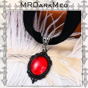 May include: A black velvet choker necklace with a decorative pendant. The pendant features a large, oval, red gemstone set in a black filigree frame. The necklace is displayed on a bed of white feathers, with the text "MRDARKMEG" at the top.