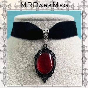 Może przedstawiać: Czarny aksamitny naszyjnik typu choker z wisiorkiem. Wisiorek ma owalny, czerwony kamień w czarnej filigranowej ramie. Nazwa marki "MRDARKMEG" jest widoczna u góry zdjęcia.