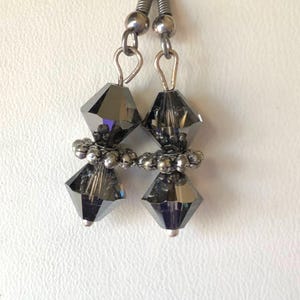 Puede incluir: Un par de pendientes colgantes con dos cuentas facetadas de color gris oscuro en forma de diamante, separadas por un pequeño espaciador redondo de metal plateado texturizado. Los pendientes están unidos a ganchos de metal plateado.