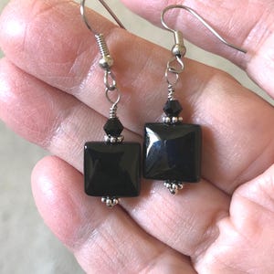 Puede incluir: Un par de pendientes cuadrados negros con ganchos y detalles plateados. Cada pendiente presenta una cuenta negra cuadrada pulida, una pequeña cuenta facetada negra y tapas de cuentas plateadas. Los pendientes se muestran en una mano abierta.