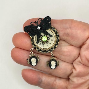May include: A steampunk-style brooch featuring a black butterfly atop a clock face with Roman numerals. The brooch has a green gem and two cameo pendants. The metal has a gold finish.