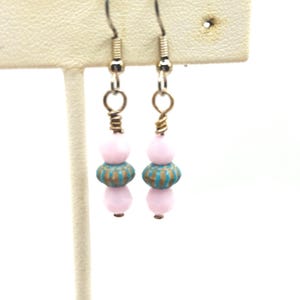 Peut inclure: Une paire de boucles d'oreilles pendantes avec des crochets argentés. Chaque boucle d'oreille comporte une perle rose clair, une perle turquoise et bronze, et une autre perle rose clair. Les boucles d'oreilles sont exposées sur un présentoir blanc.