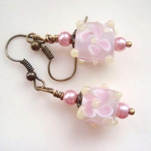 Può includere: Un paio di orecchini fatti a mano con perline a cubo floreali rosa, perline di perle rosa e componenti metallici color bronzo antico. Gli orecchini hanno una chiusura a gancio e sono per orecchie forate.