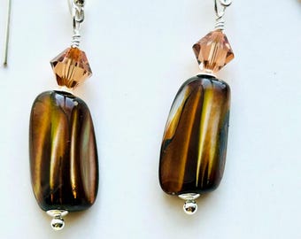 May include: A pair of handcrafted earrings featuring iridescent brown and gold oval beads. Each earring has a small, faceted peach-colored crystal and a silver-tone hook. The earrings are designed for pierced ears and are displayed against a white background.
