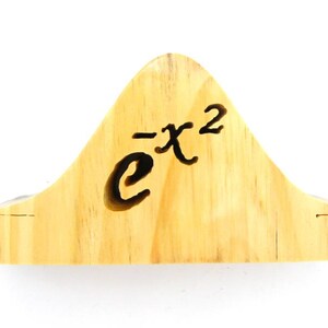 May include: A wooden sculpture of the mathematical function e to the power of negative x squared. The sculpture is carved from light brown wood and has a smooth, rounded surface.