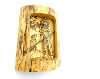 May include: A wood carving of a person with a dog. The carving is set in a wooden frame. The person is wearing a hat and holding a stick. The dog is standing next to the person.