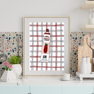 May include: Framed print featuring a tube of tomato paste against a red, gray, and white plaid background. The tube is labeled "Double Concentrated Tomato Paste" with a drawing of a tomato. The artwork is displayed in a kitchen setting.