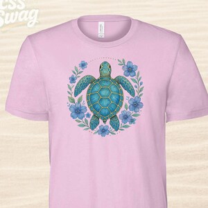 Puede incluir: Camiseta rosa claro con un dise&ntilde;o de tortuga marina turquesa rodeada de flores azules y hojas verdes. La tortuga tiene un patr&oacute;n de caparaz&oacute;n detallado. El texto "CSS Swag" est&aacute; en la esquina superior izquierda.