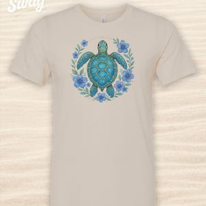 Puede incluir: Camiseta color crema con un dise&ntilde;o detallado de tortuga marina turquesa rodeada de flores azules y hojas verdes. La tortuga tiene un caparaz&oacute;n estampado. La camiseta tiene mangas cortas y cuello redondo.