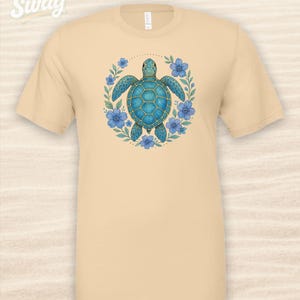 Puede incluir: Una camiseta beige con un dise&ntilde;o de tortuga marina azul rodeada de flores azules y hojas verdes. La camiseta tiene el texto "CSS Swag" en la esquina superior izquierda.