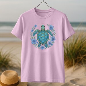 Puede incluir: Camiseta rosa claro con un dise&ntilde;o de tortuga marina turquesa rodeada de flores azules. La tortuga tiene un patr&oacute;n de caparaz&oacute;n detallado. La camiseta est&aacute; colgada en una percha sobre un fondo de playa.