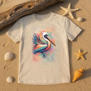 May include: A cream-colored t-shirt featuring a colorful watercolor illustration of a pelican. The bird has a long beak and is surrounded by splashes of pink, blue, and yellow. The shirt is displayed on a sandy surface with seashells and starfish.