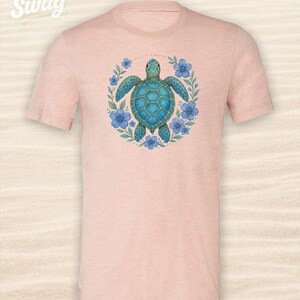 Puede incluir: Camiseta rosa claro con un dise&ntilde;o de tortuga marina turquesa rodeada de flores azules y hojas verdes. El texto "CSS Swag" es visible en la parte superior izquierda. Cuello redondo.