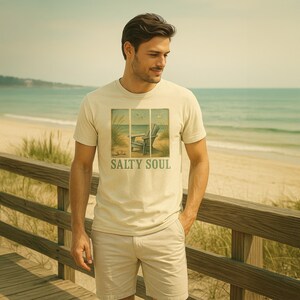 May include: A light beige t-shirt with the text "SALTY SOUL" and a beach scene graphic. The graphic features a wooden chair on a sandy beach with tall grass and birds. The person is wearing khaki shorts and standing on a wooden boardwalk.