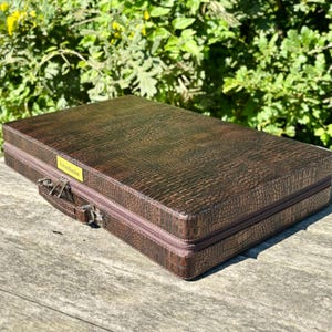 A brown leather briefcase with a crocodile skin texture sits on a wooden surface, with a lush green background.