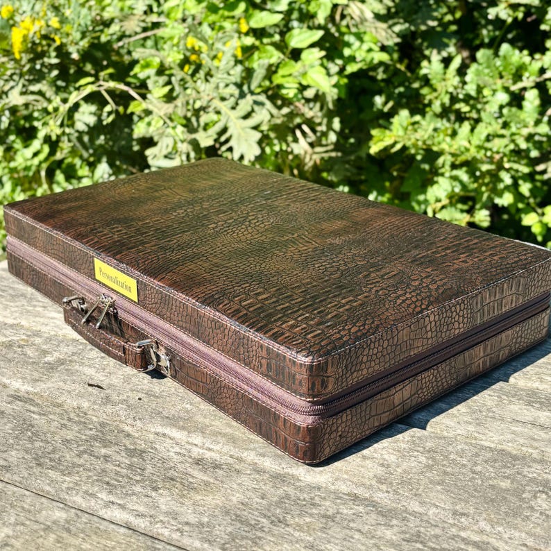 A brown leather briefcase with a crocodile skin texture sits on a wooden surface, with a lush green background.