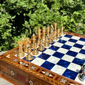 A chess set with golden pieces is placed on a blue and white checkered board, with a natural outdoor background.