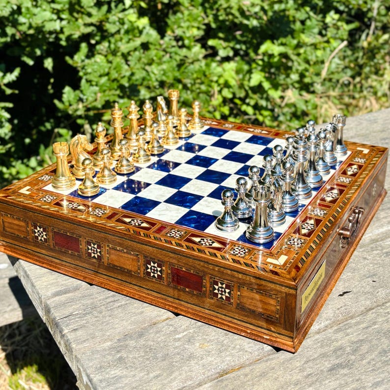 A chess set with a blue and white checkered board and gold pieces is displayed on a wooden surface, with a lush green background.