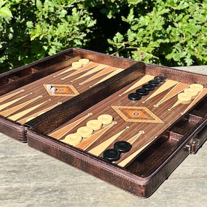 May include: A brown leather backgammon set, open to reveal the playing surface. The board features a wood grain design with light and dark triangles, diamond-shaped inlays, and black and white game pieces. Dice are visible.