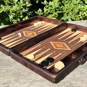 An open brown leather game case containing a backgammon game board and pieces, placed on a wooden surface with greenery in the background.