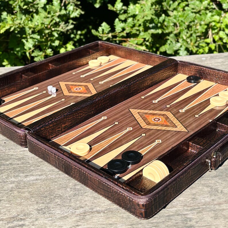 An open brown leather game case containing a backgammon game board and pieces, placed on a wooden surface with greenery in the background.