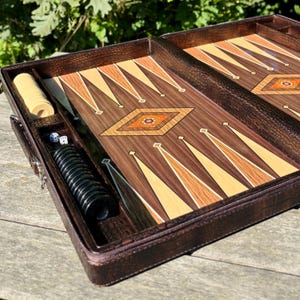 A brown leather-bound game board with a decorative pattern sits on a wooden surface, accompanied by a set of dice and a game piece.